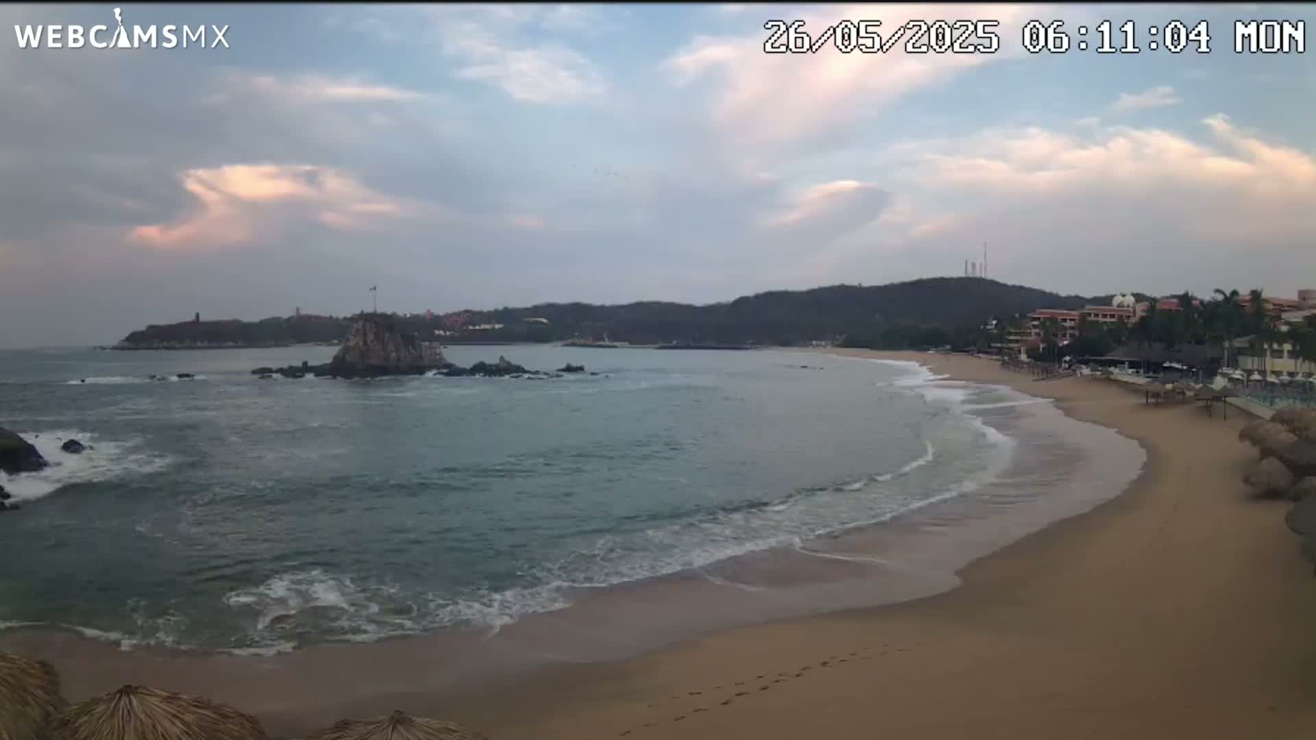 A sandy beach curves gently into a calm ocean under a partly cloudy sky, with a rocky island and distant coastline visible.