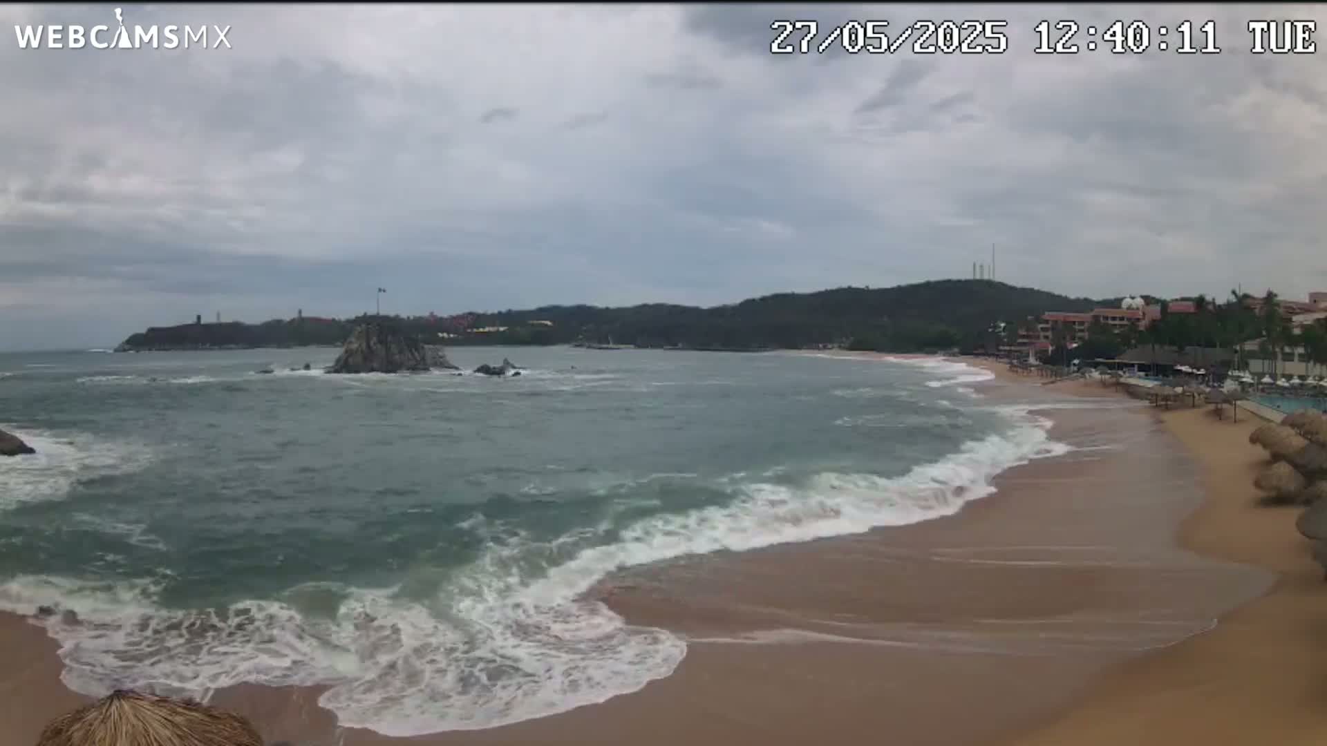 A sandy beach with moderate waves under an overcast sky meets a rocky outcrop in a bay bordered by hills and buildings.