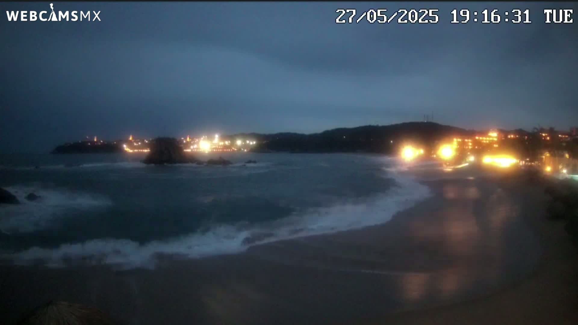 A dark, nighttime beach scene shows waves crashing on the shore near a town with lights illuminating the coastline under a mostly cloudy sky.
