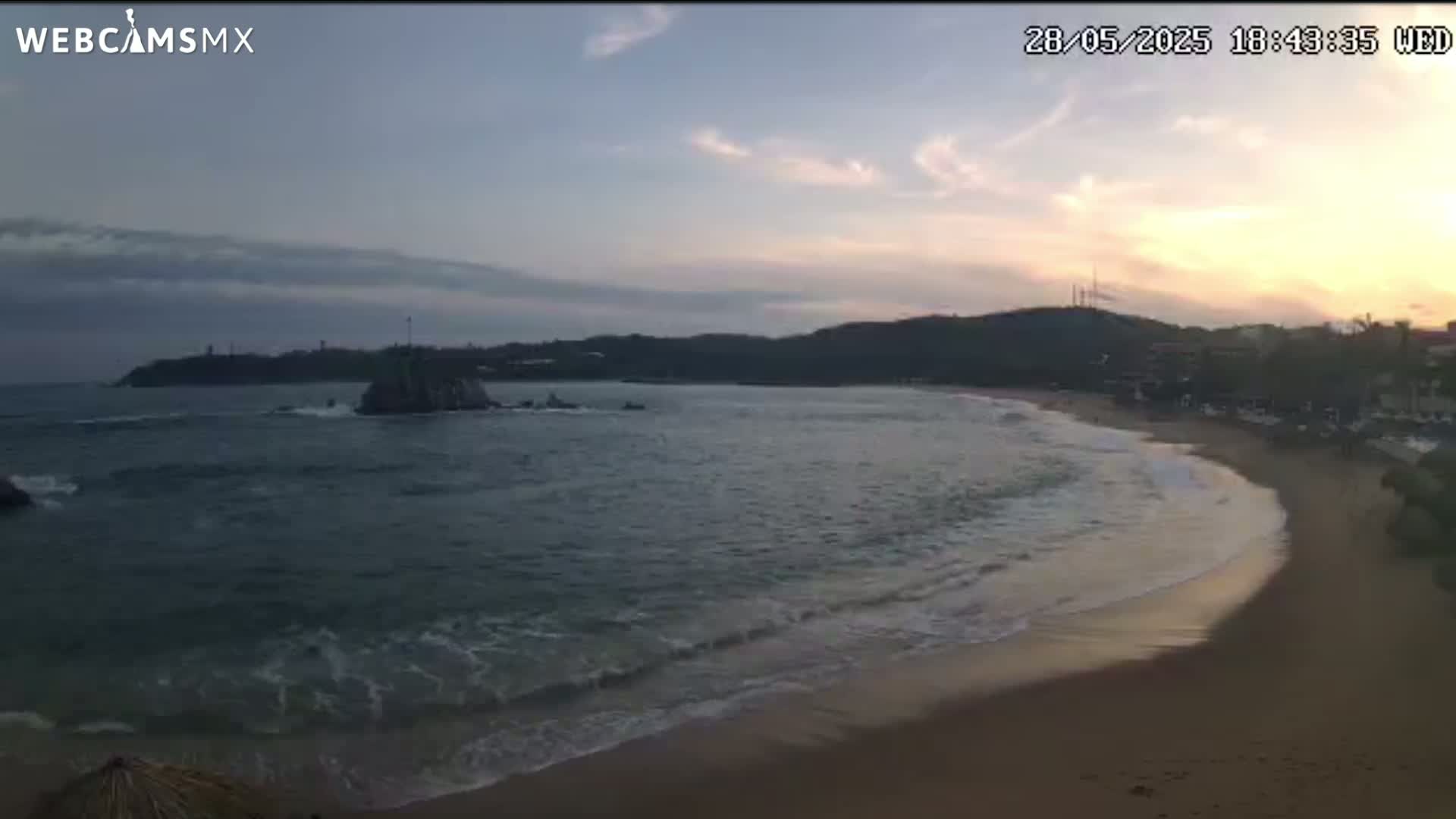 A sandy beach curves gently around a calm ocean at sunset, with dark hills rising in the background under a partly cloudy sky.