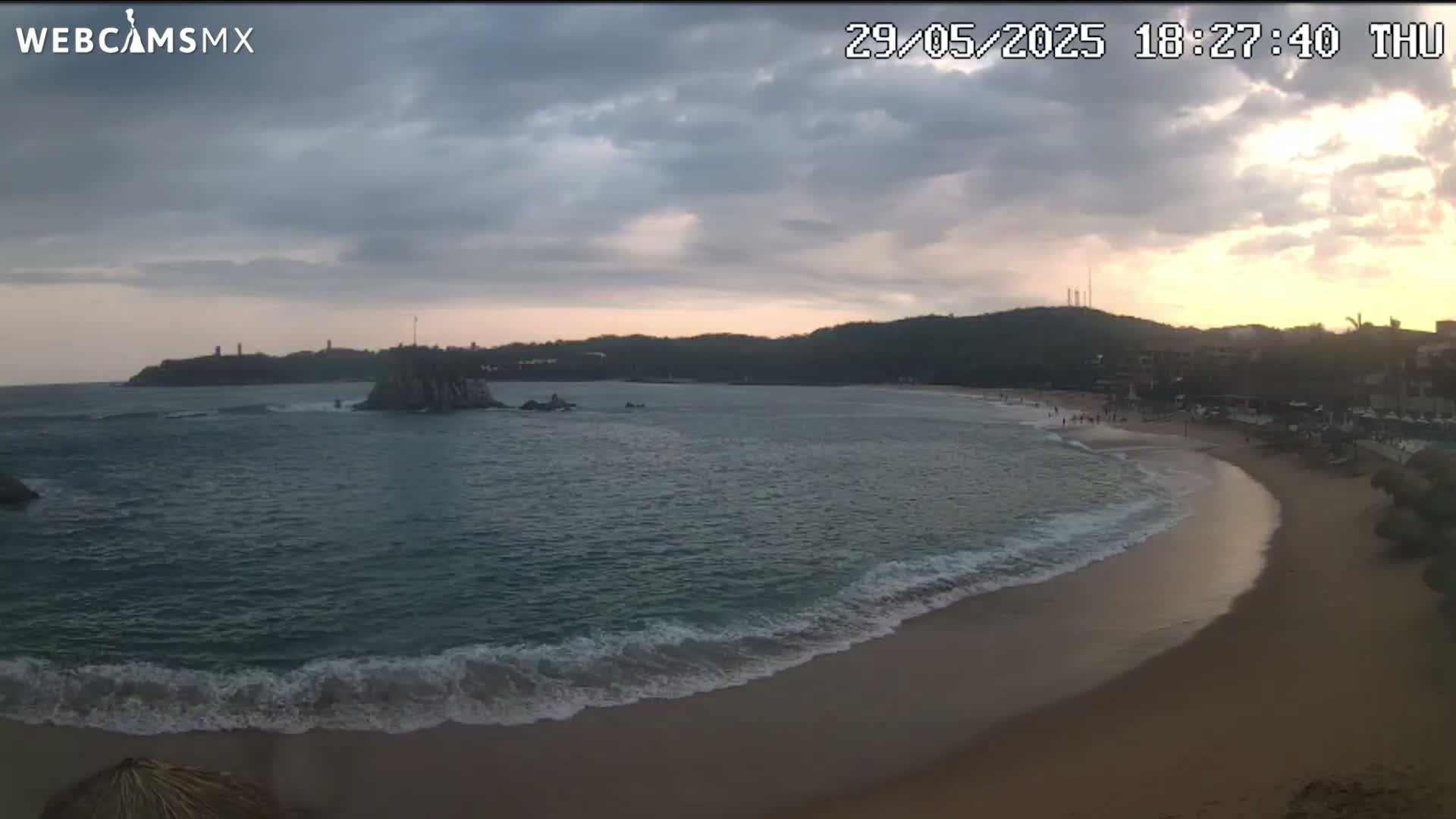 A sandy beach curves around a calm bay under a partly cloudy sunset sky.