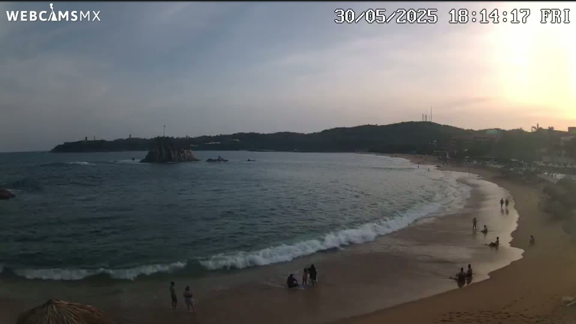 A beach scene at sunset shows waves gently lapping the shore, with a few people scattered on the sand and in the water under a mostly clear sky.