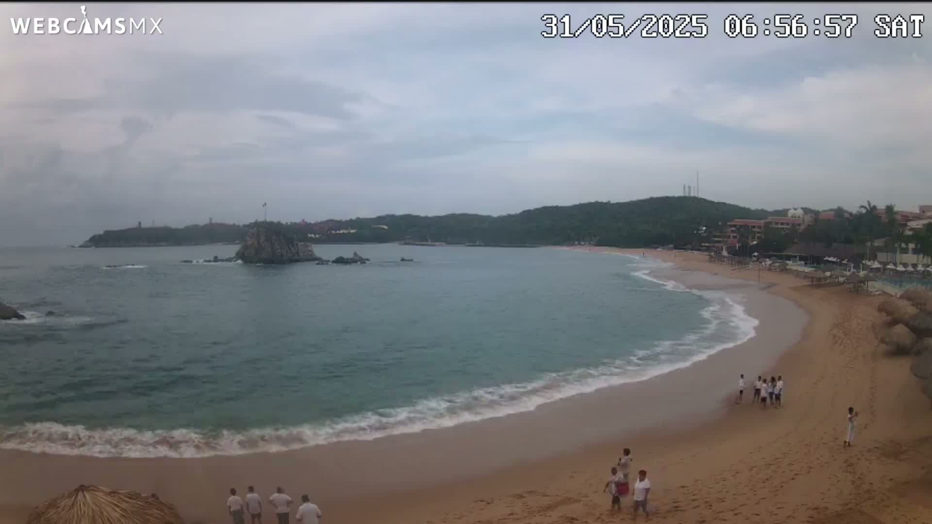 A sandy beach curves into a calm, grey-sky ocean, with a rocky island and distant buildings visible.