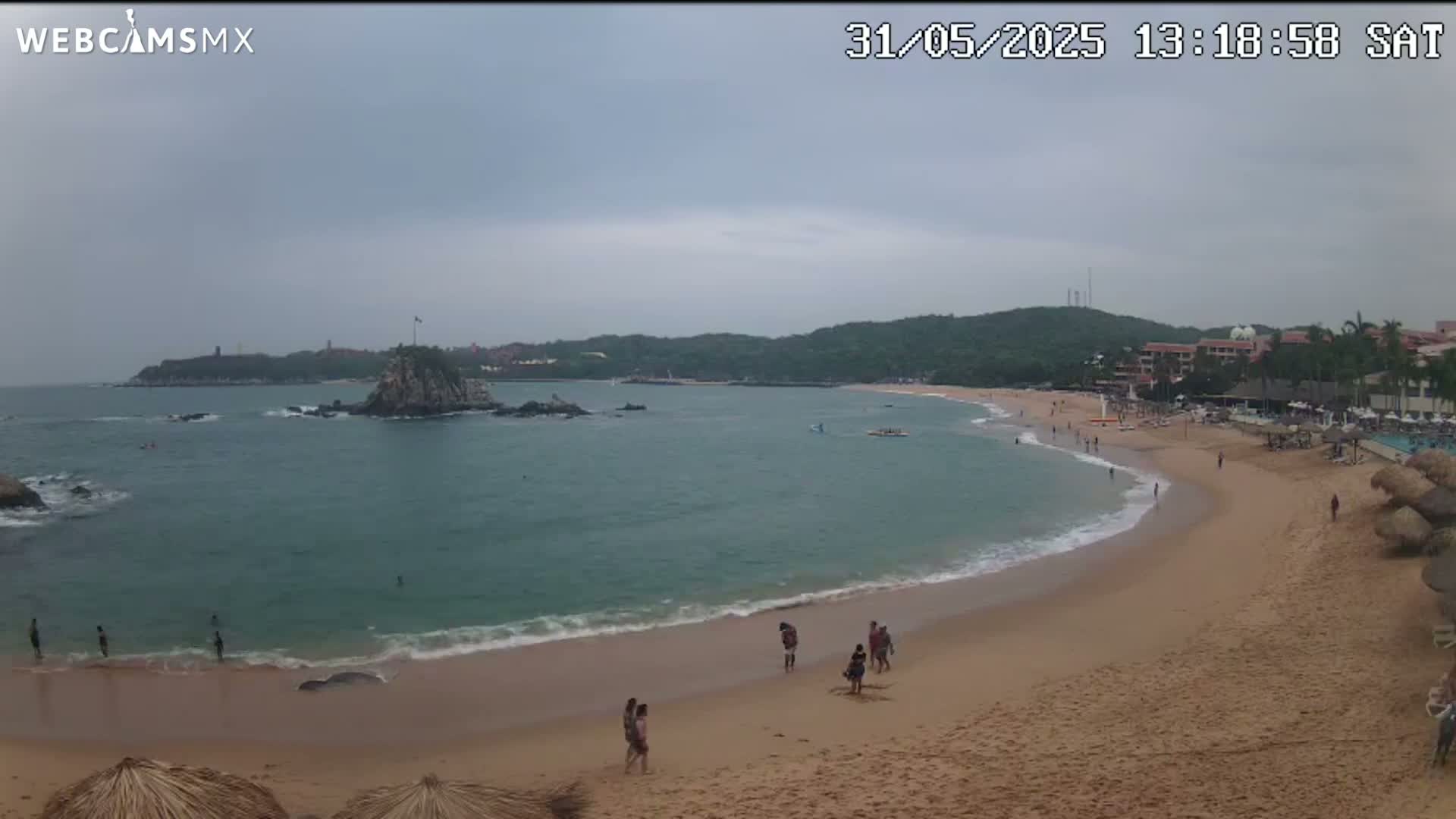 A sandy beach curves around a calm bay under an overcast sky, with a rocky island and distant green hills visible.