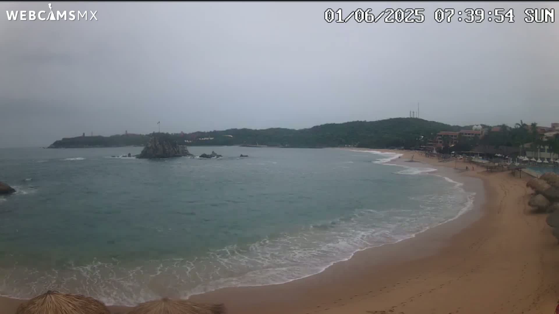 A mostly cloudy day shows a sandy beach with gentle waves lapping the shore, a rocky outcropping in the calm water, and a distant hilly coastline.