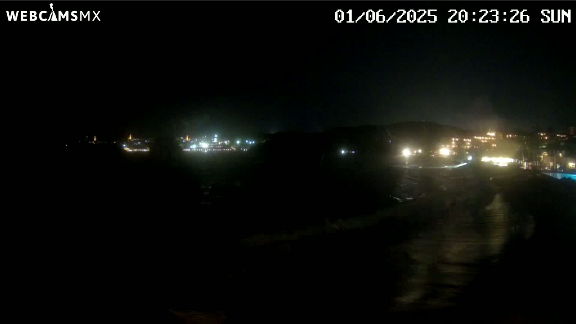 A nighttime, dark view of a coastal town with lights visible on a hillside and along the water.