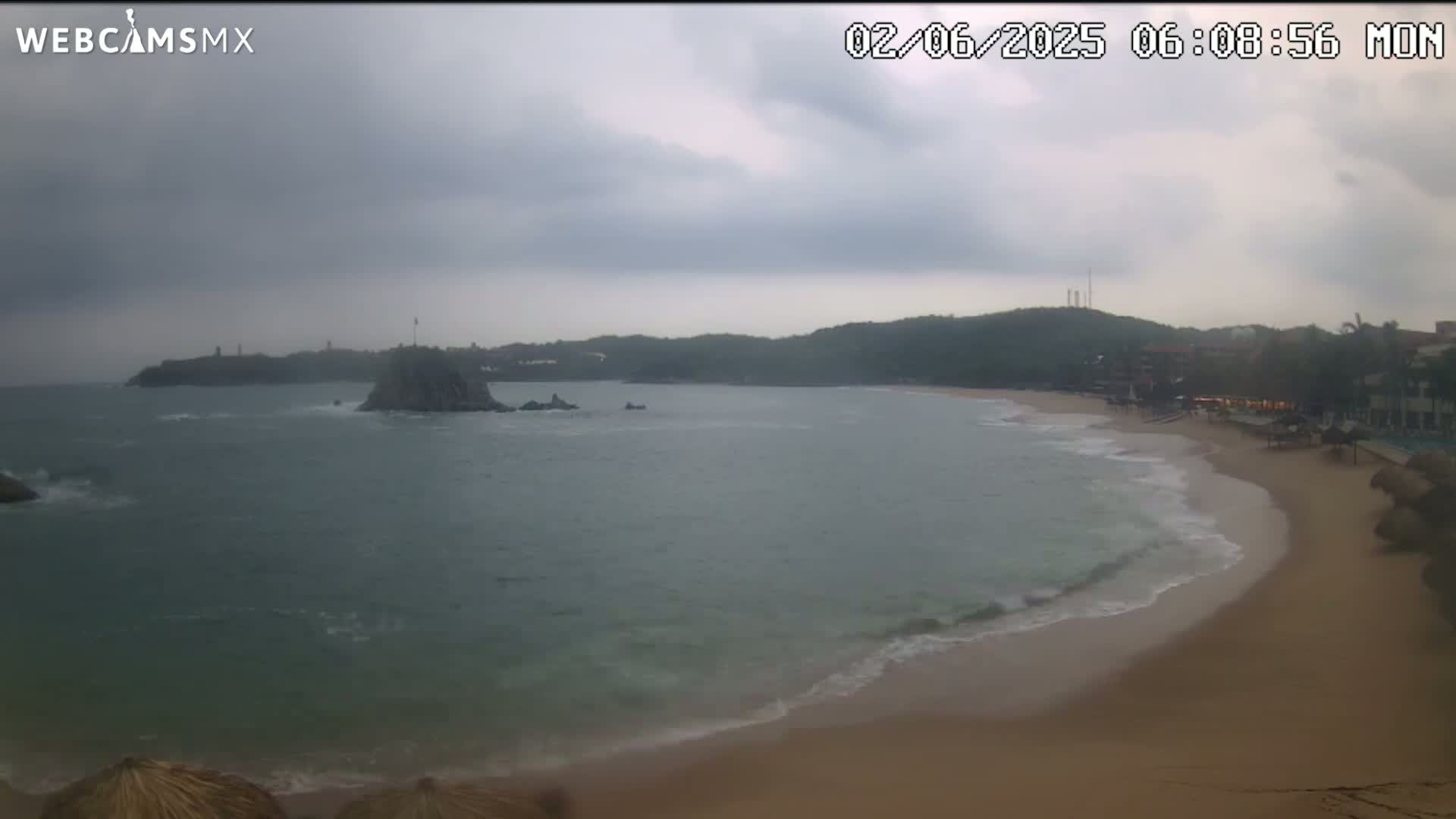 A sandy beach curves into a calm ocean under an overcast sky, with a rocky island and distant hills visible.