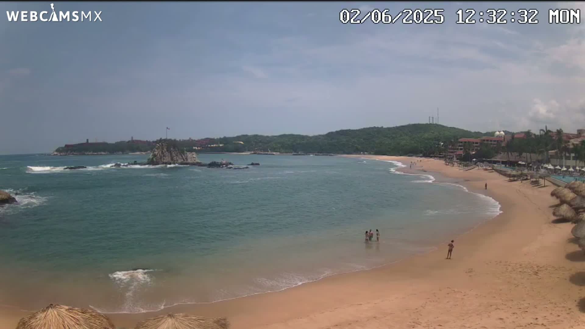 A mostly sunny day at a beach with calm, clear water, a sandy shore, and a few people swimming and walking along the sand, backed by green hills and buildings.