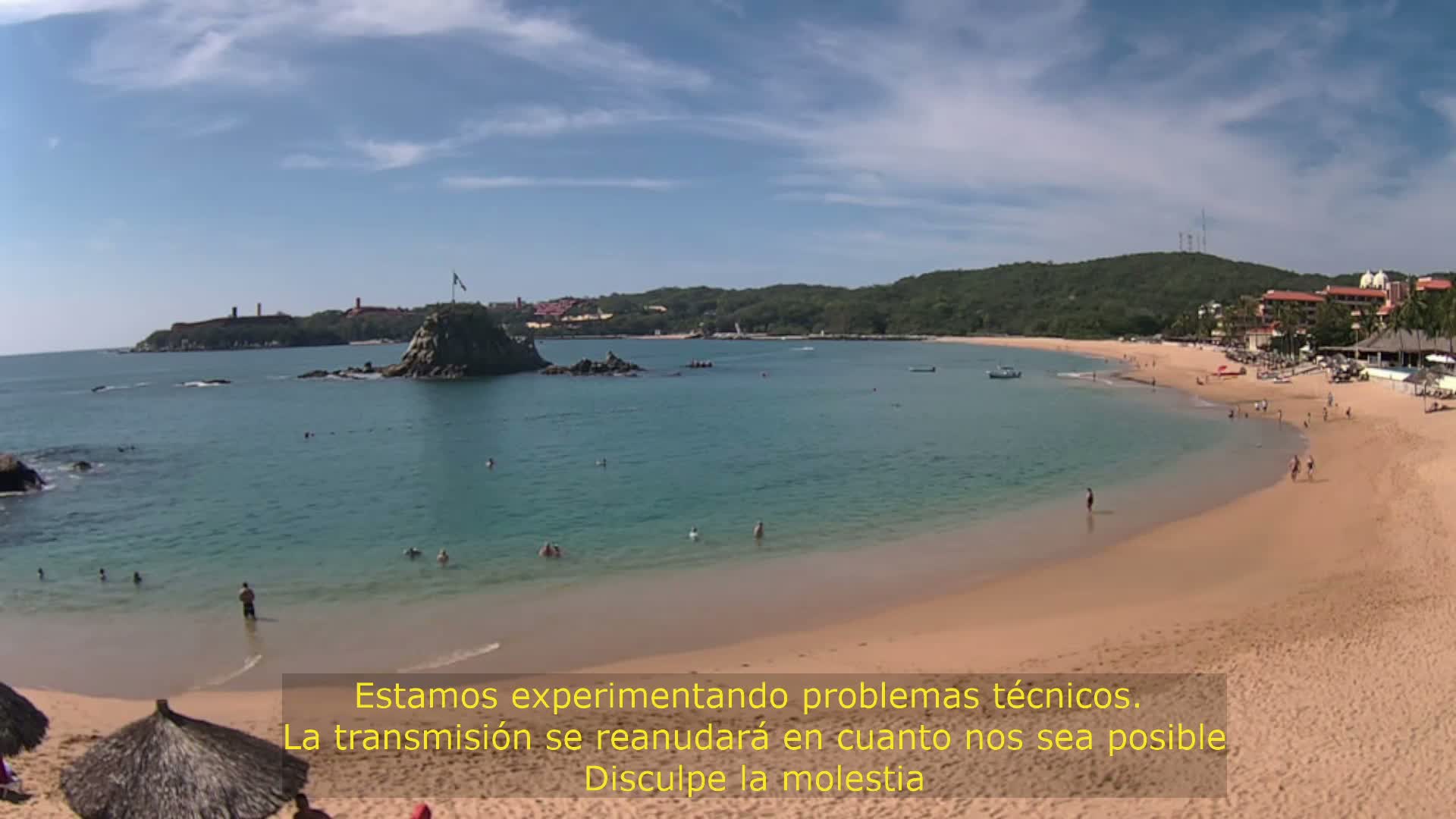 A calm, sunny day shows a secluded bay with a sandy beach and calm, clear water, with a few people swimming and walking along the shore.