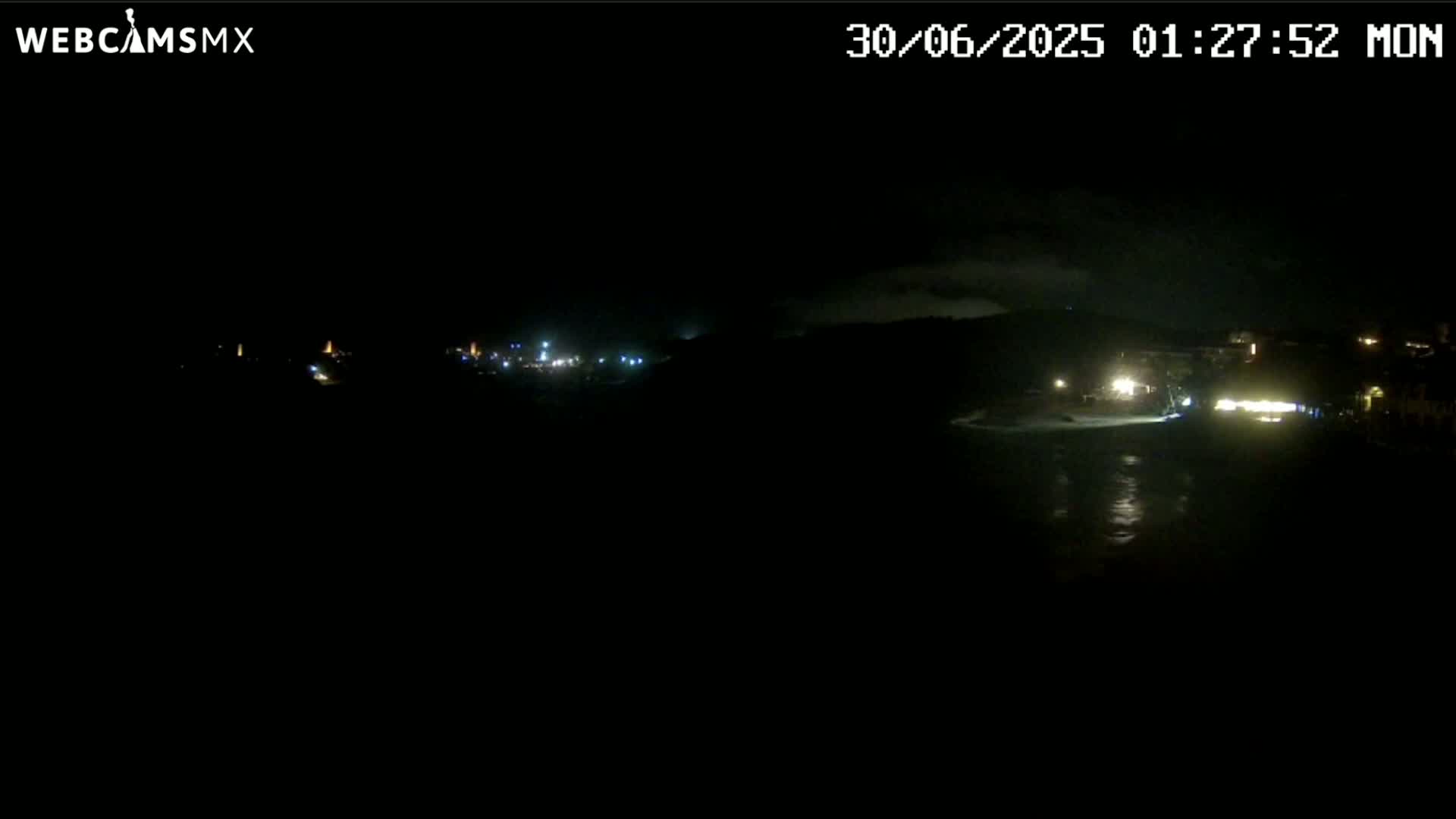 A nighttime view of a dark coastline with a few faint lights visible in the distance under a cloudy sky.