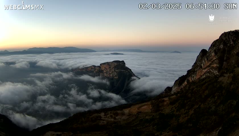 A mountaintop view at sunrise shows a vast expanse of clouds below, with other mountains and hills in the distance.