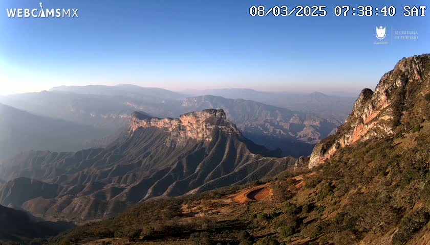 A high-angle, long shot shows a vast, mountainous landscape under a clear blue sky, with hazy, distant mountains visible.