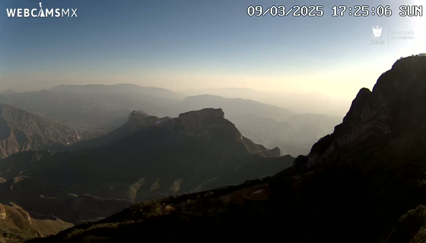 A hazy sunset illuminates a vast, mountainous landscape, with dark, rugged peaks silhouetted against the fading light.