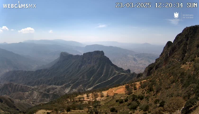 A hazy, sunny day reveals a vast, mountainous landscape with deep valleys and rugged, dark-colored peaks.