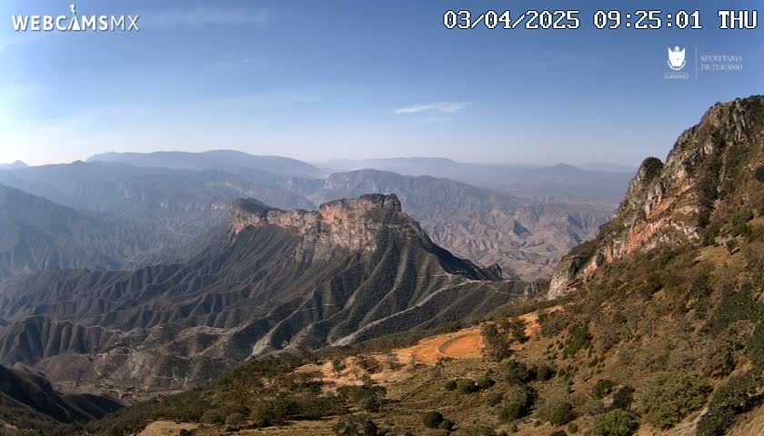 A vast, mountainous landscape under a clear blue sky stretches to the horizon, showing a variety of rock formations and vegetation.