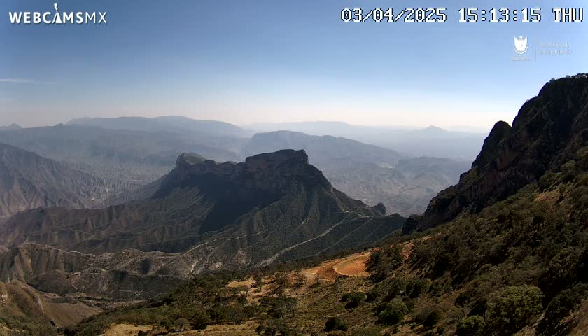 A high-angle view shows a vast, mountainous landscape under a clear, sunny sky.