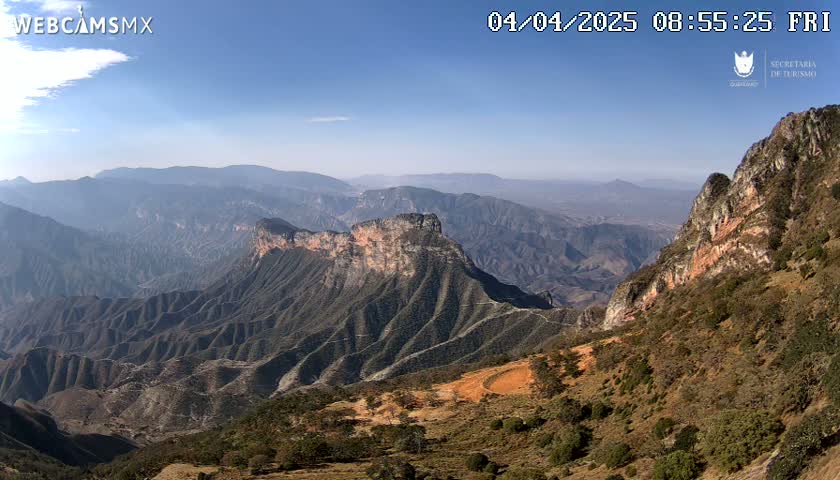 A vast, mountainous landscape stretches under a clear, sunny sky, revealing layers of rugged, eroded peaks and valleys.