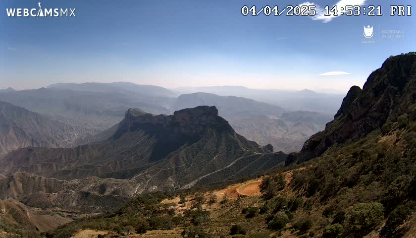 A high-angle view shows a vast, mountainous landscape under a clear, bright sky, with sparse vegetation covering the hillsides.