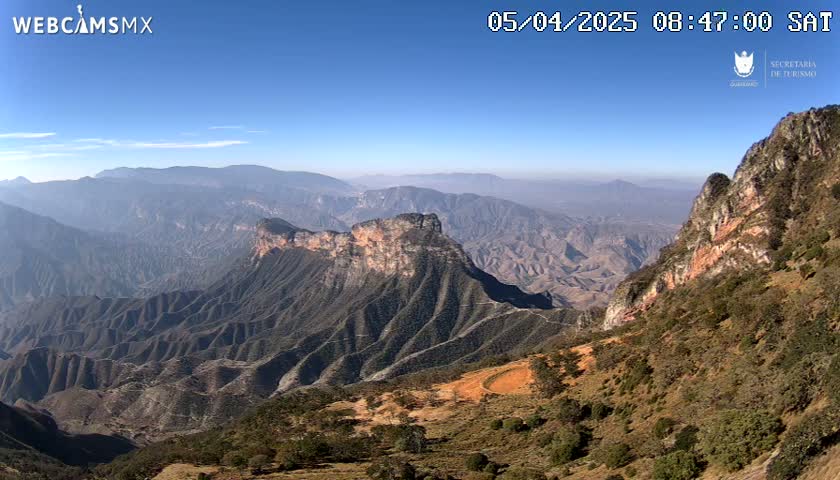 A vast, mountainous landscape under a clear blue sky displays a rocky, layered terrain with varying shades of brown and green vegetation.