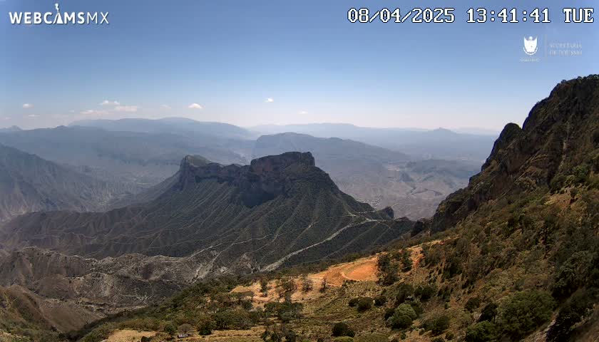 A high-angle view on a clear day shows a vast, mountainous landscape with deep canyons and valleys, exhibiting a mix of rocky terrain and sparse vegetation.