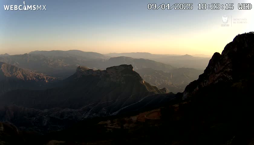 A panoramic view of a vast, mountainous landscape during sunset, with hazy atmospheric conditions.