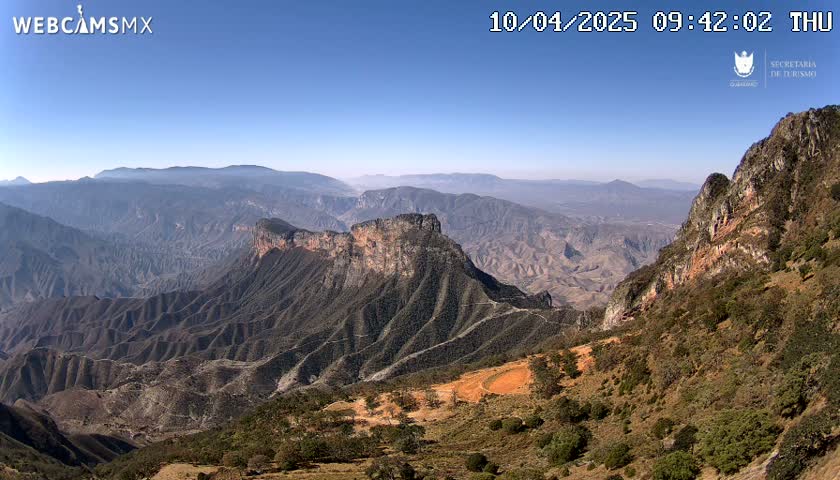 A vast, mountainous landscape under a clear blue sky stretches to the horizon, revealing layers of rocky terrain and sparse vegetation.