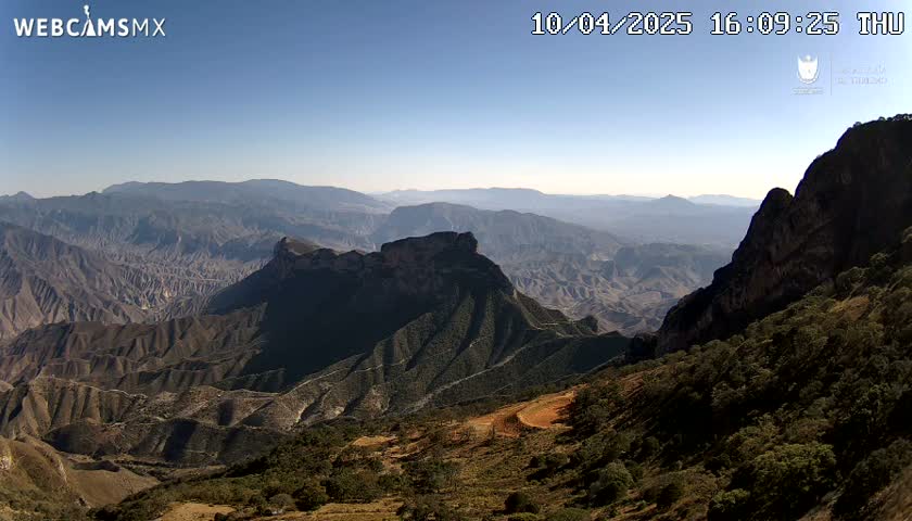 A vast, mountainous landscape under a clear blue sky stretches into the distance, showing various shades of brown and green.