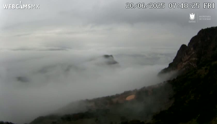 A rocky mountainside is shrouded in a thick fog, with a valley below partially obscured by the dense clouds.