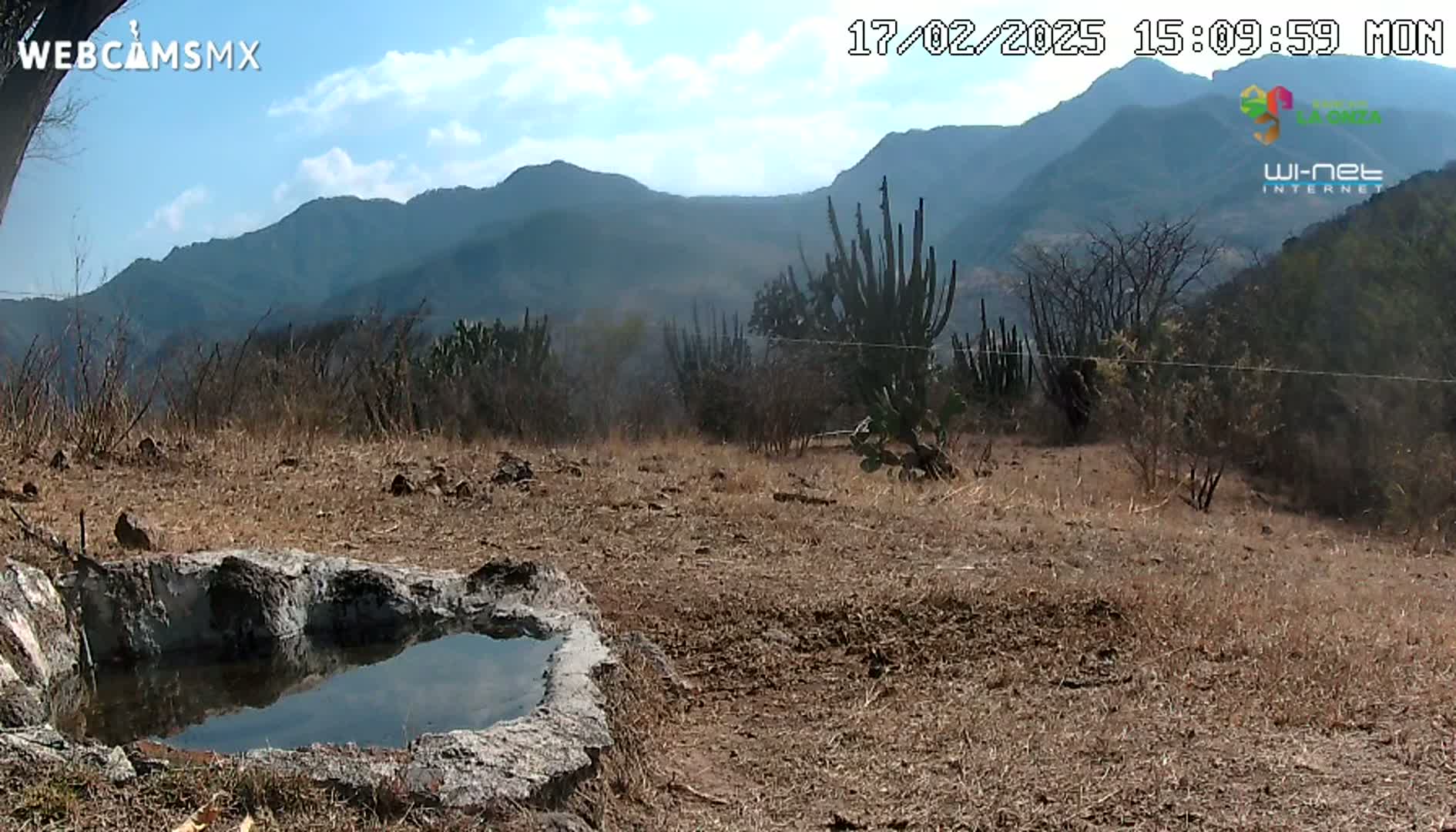 A small, stone-lined pool of water sits in a dry, brown landscape against a backdrop of distant, hazy mountains under a partly cloudy sky.