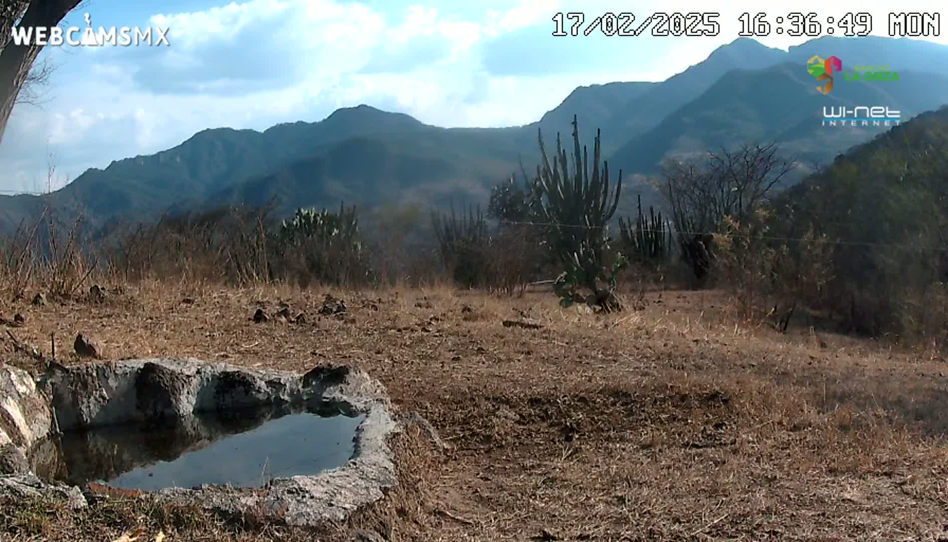 A stone basin containing still water sits in a dry, brown landscape against a backdrop of distant mountains under a partly cloudy sky.