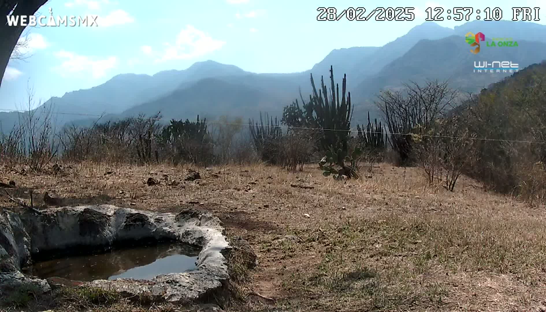 A small stone basin containing still water sits in a dry, brown landscape under a partly cloudy sky, with mountains in the background.