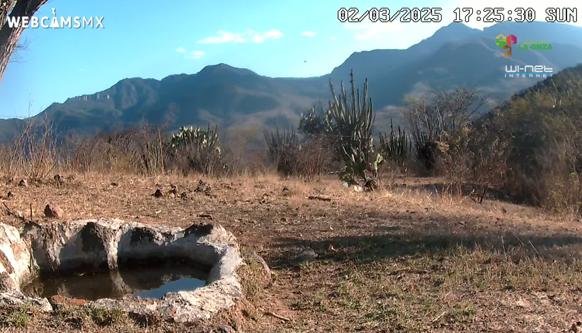 A stone water trough sits in a dry, brown landscape under a mostly sunny sky, with mountains in the background.