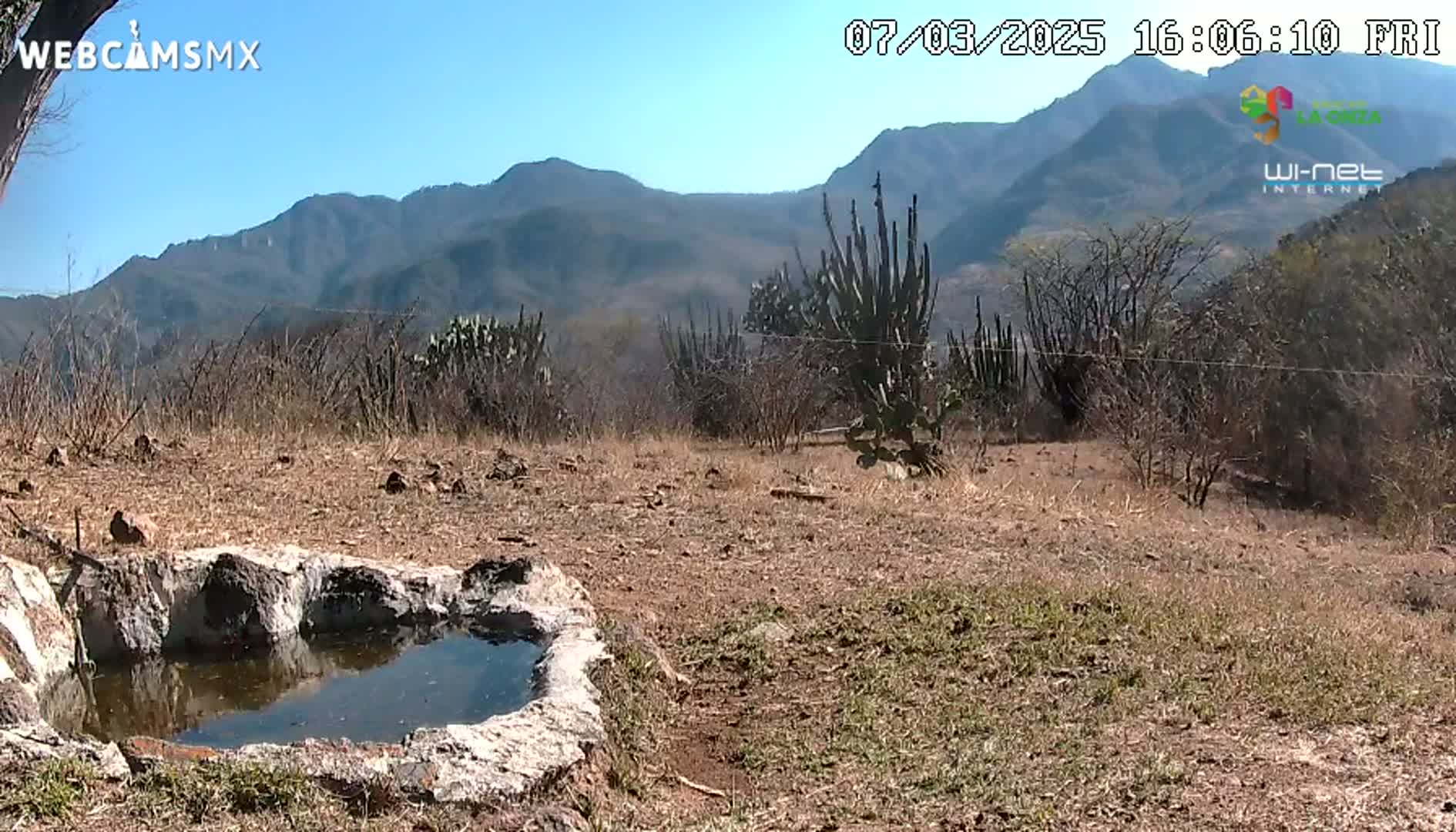 A small, stone-lined pool of water sits in a dry, sparsely vegetated landscape under a clear blue sky, with mountains in the background.