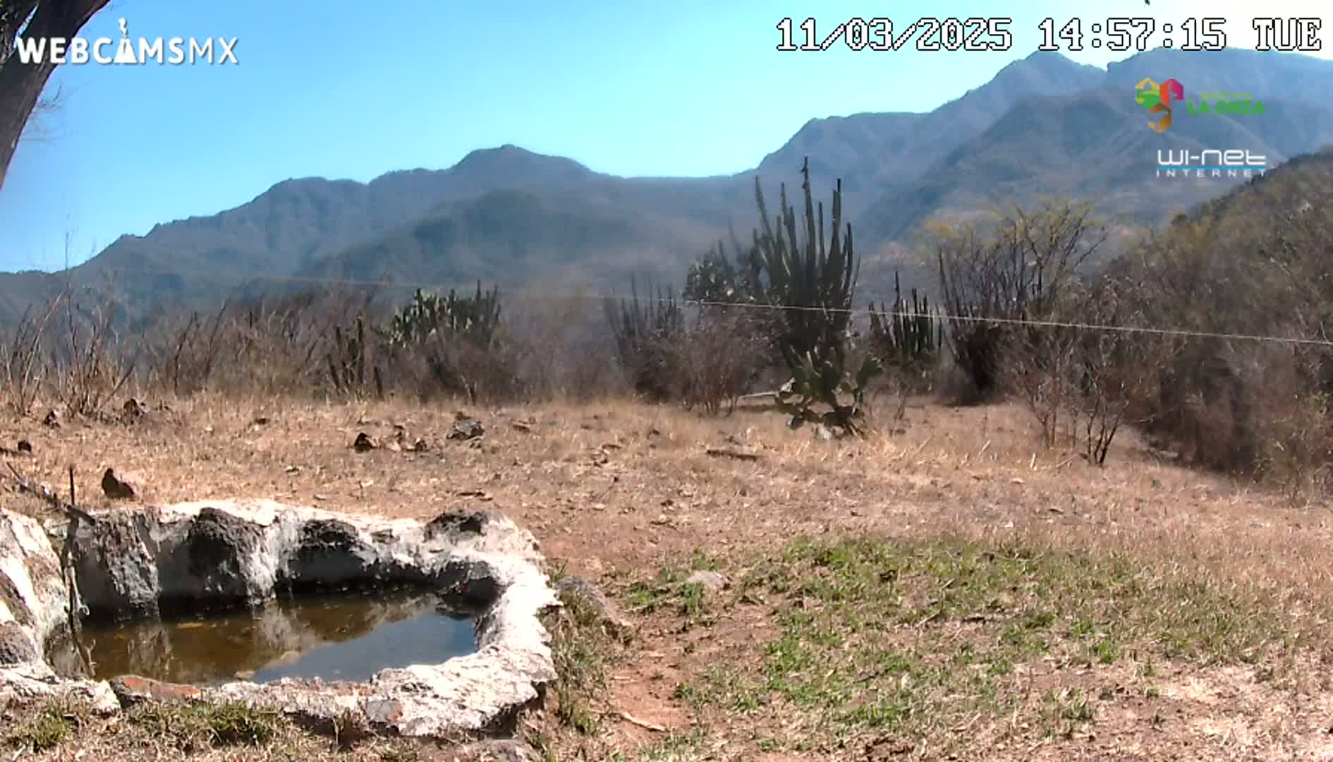 A small, stone-lined water basin sits in a dry, brown landscape under a clear blue sky, with mountains visible in the distance.