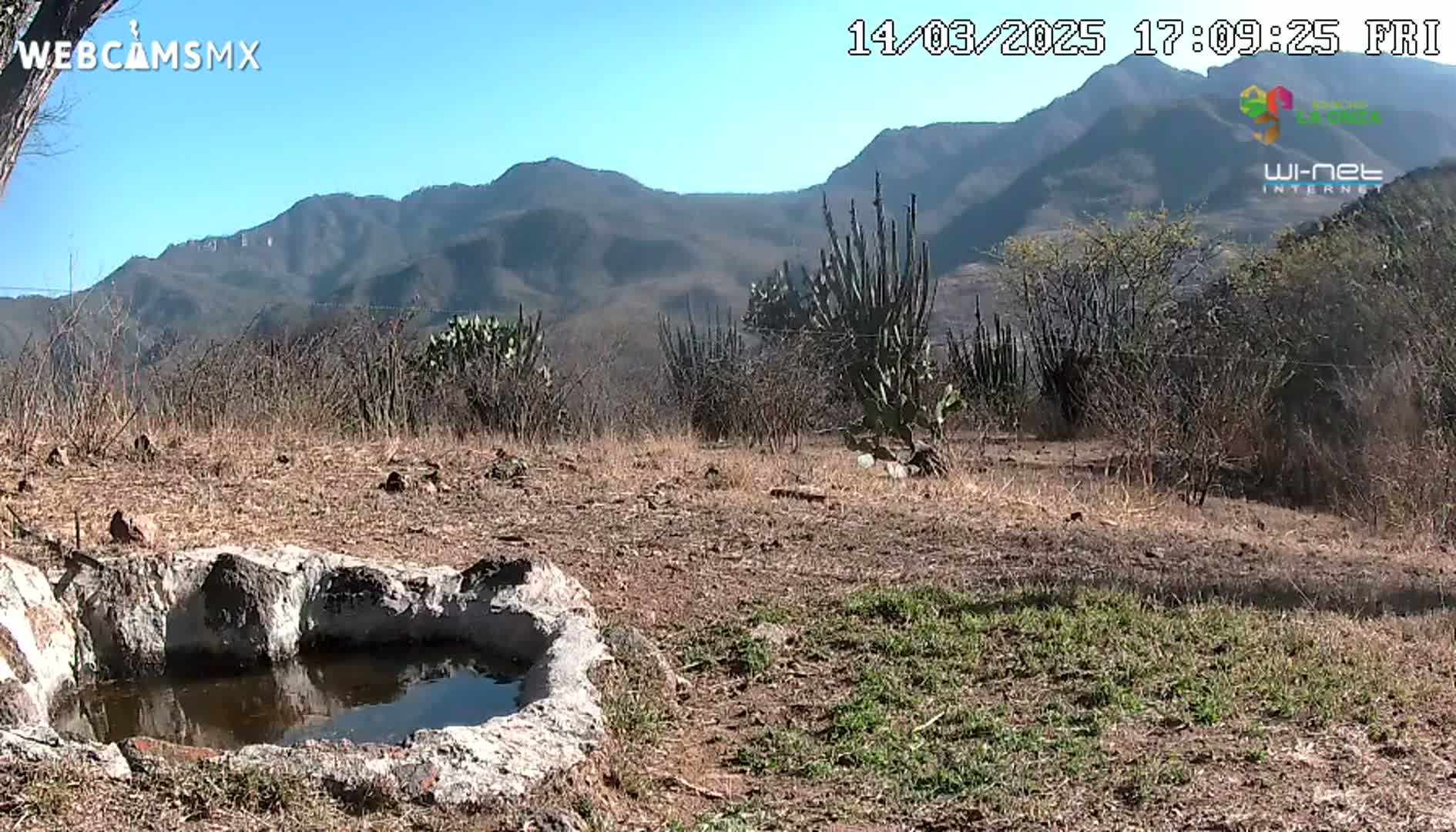 A small, stone-lined pool of water sits in the foreground of a dry, scrubby landscape under a clear blue sky, with mountains visible in the background.