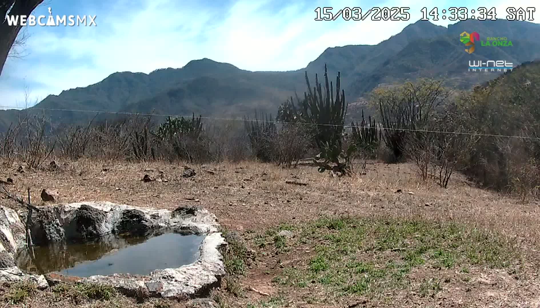 A small, stone-lined pool of water sits in a dry, scrubby landscape under a partly cloudy sky, with mountains in the background.
