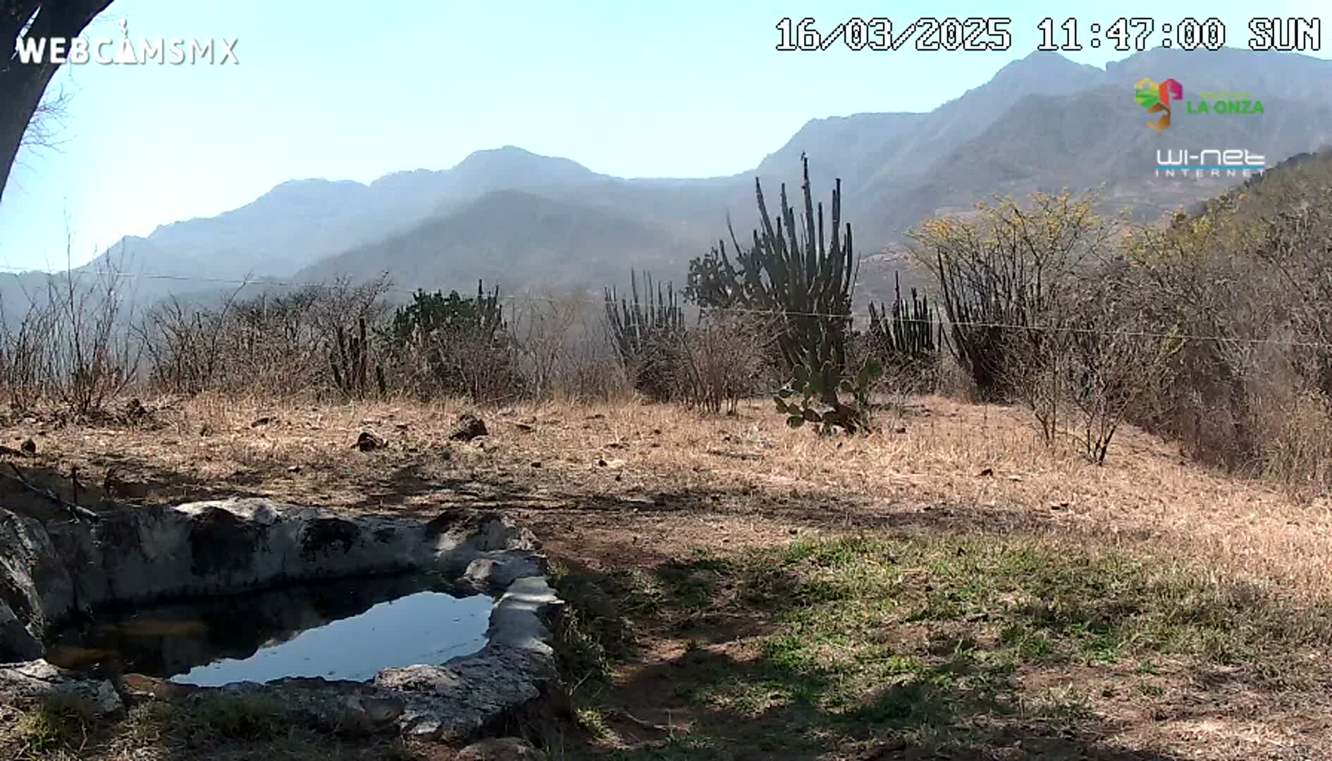 A small, stone-lined pool of water sits in a dry, scrubby landscape under a sunny sky, with mountains visible in the distance.