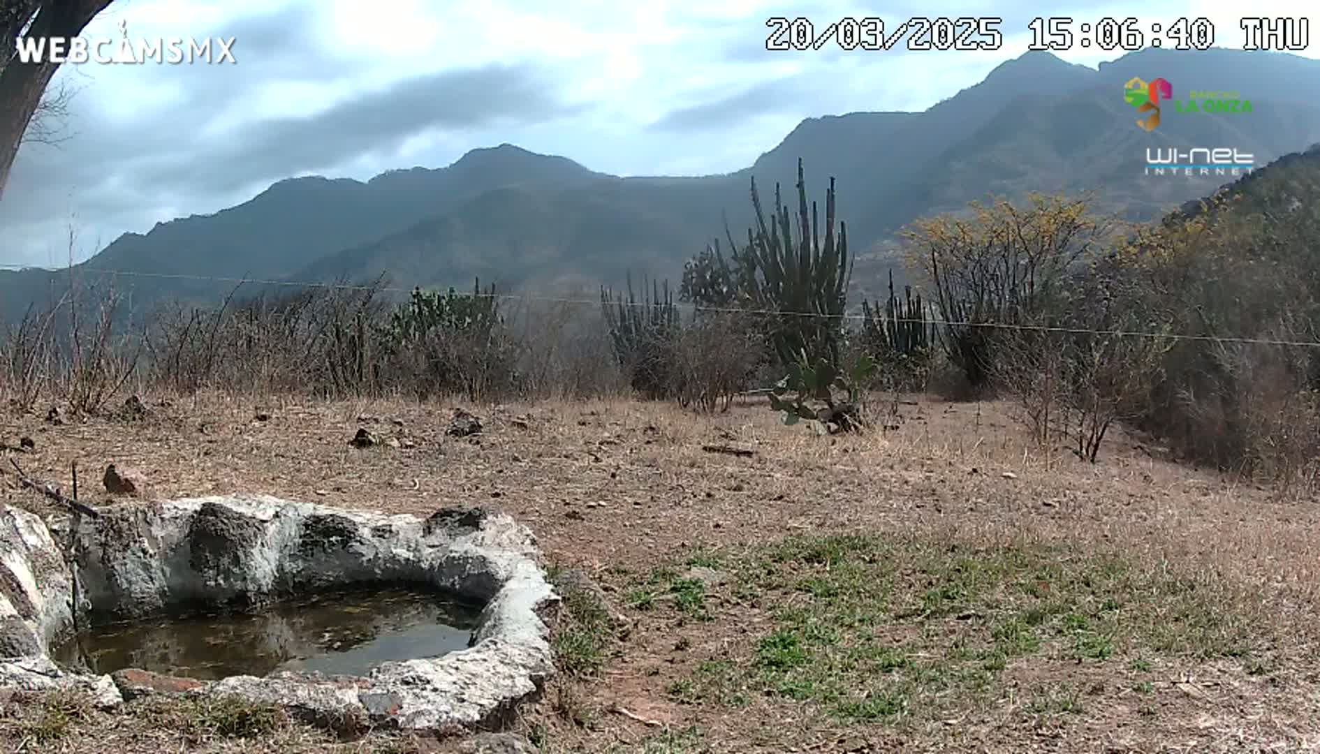A stone basin containing still water sits in a dry, sparsely vegetated landscape under a partly cloudy sky, with mountains in the background.