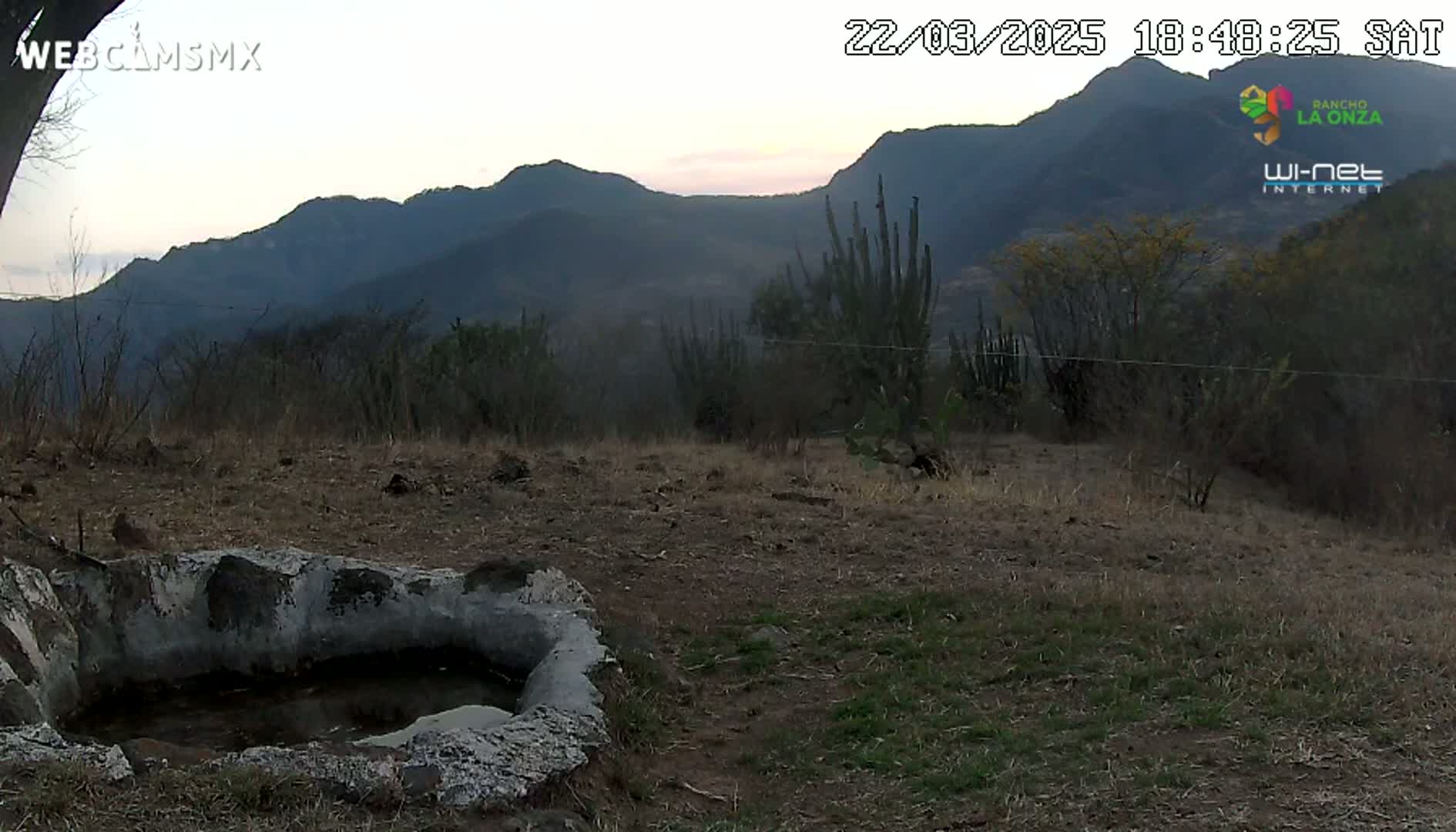 A low-lying, stone-lined water trough sits in the foreground of a dry, scrubby landscape under a twilight sky, with dark mountains in the background.