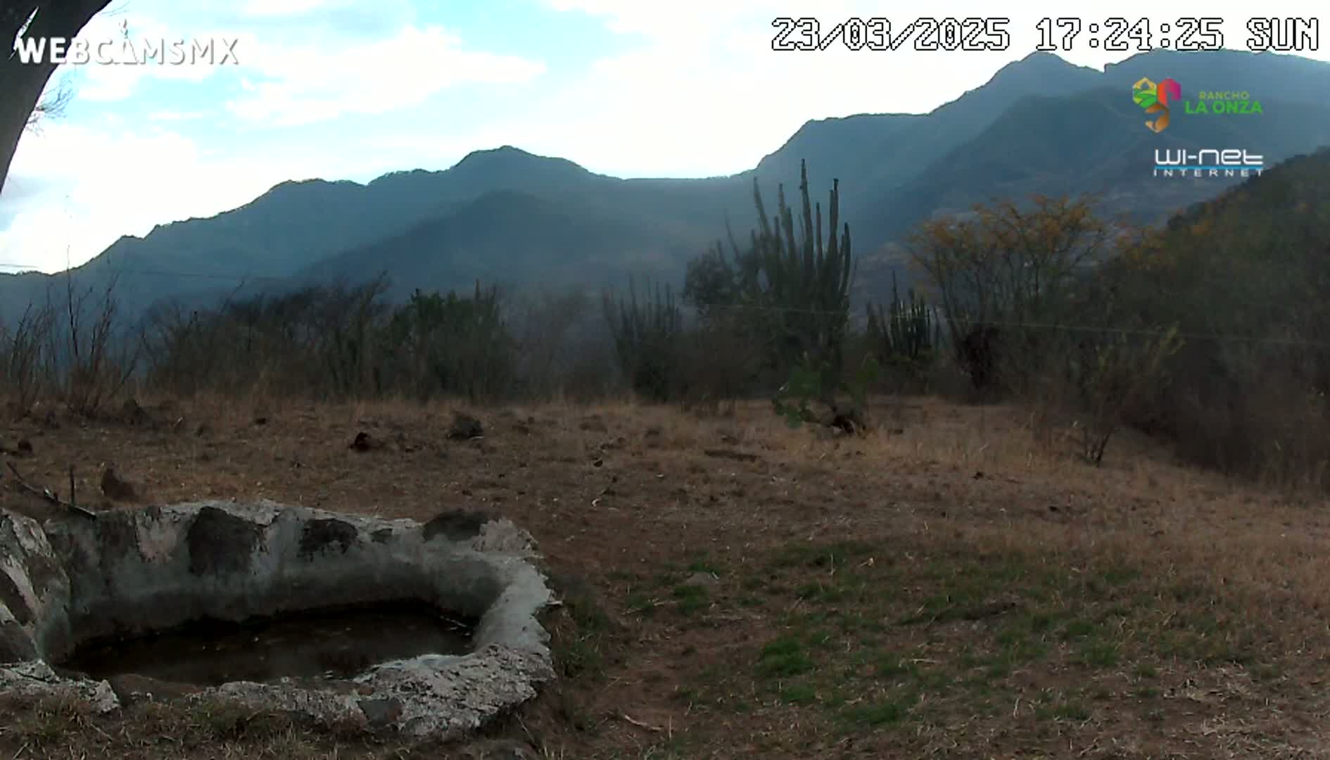 A partially-filled, cement water basin sits in a dry, scrubby landscape under a partly cloudy sky, with mountains in the background.