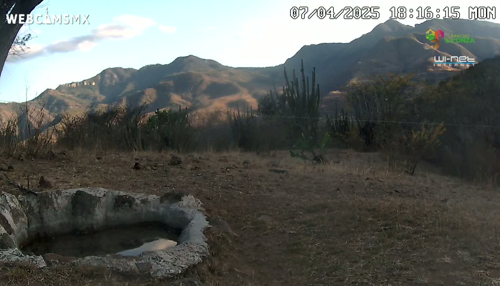 A partly cloudy day reveals a rocky, mountainous landscape with scrubby vegetation surrounding a small, stone-lined water basin.