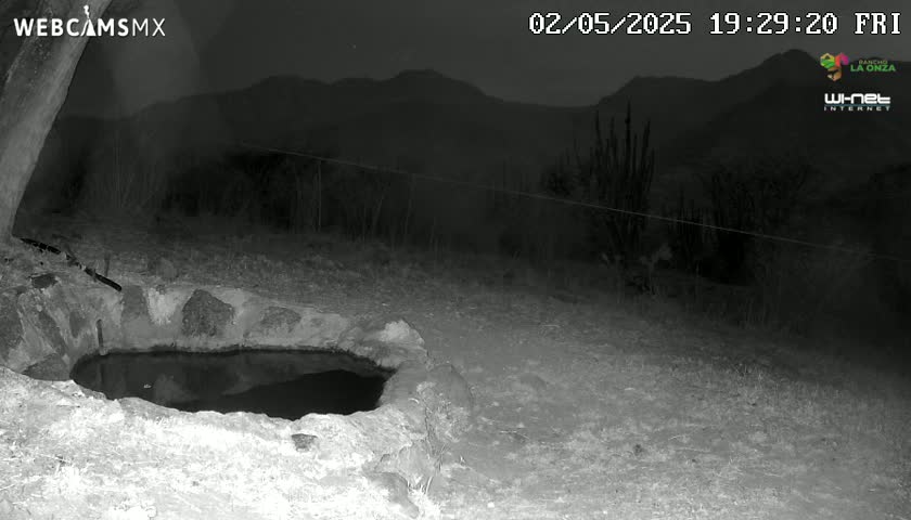 A nighttime, low-light image shows a stone-lined pool of water in the foreground, with a wire fence and dark mountains in the background under a clear sky.
