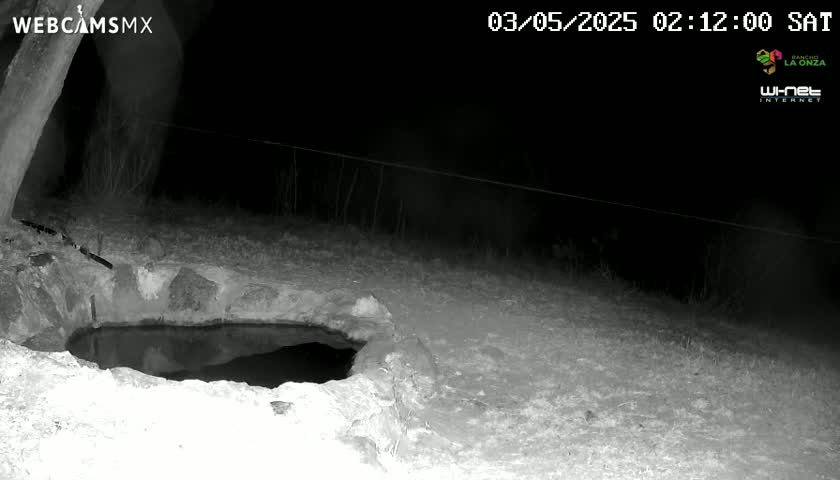 A nighttime, low-light image shows a small, dark pool of water nestled within a rocky, snow-covered area.