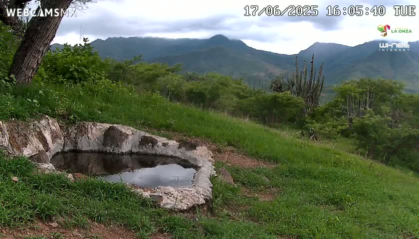 A stone basin containing still water sits in a grassy field overlooking a mountain range under a cloudy sky.