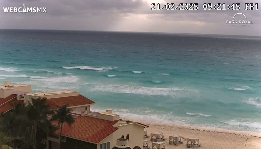 A mostly cloudy day shows turquoise ocean waves breaking on a sandy beach with several cabanas and buildings with red tile roofs visible in the foreground.