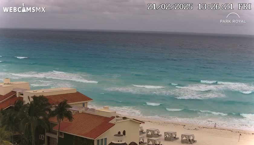 A partly cloudy day shows turquoise water with small waves lapping a sandy beach, where several beach cabanas are set up in front of a low-rise building with a red tile roof.
