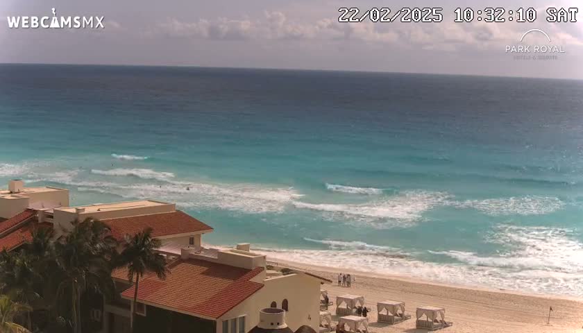 A mostly sunny day at a beach with turquoise water and small waves, showing a sandy shore and a red-roofed building partially obscured by palm trees.