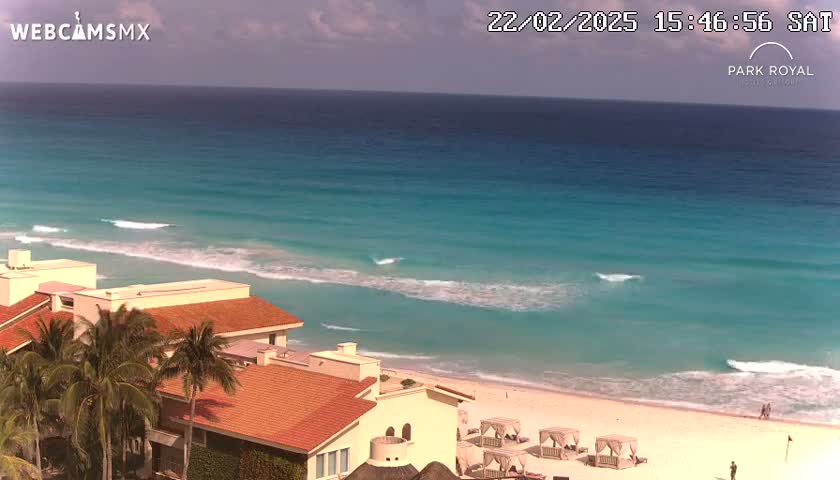 An elevated view shows a beach with turquoise water and small waves lapping a sandy shore, with several low buildings and palm trees visible in the foreground under a mostly clear sky.