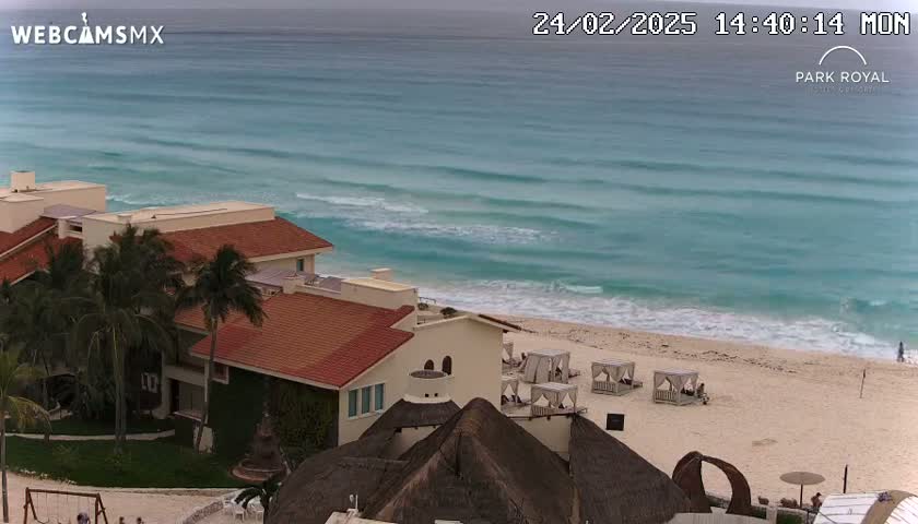 A beachfront resort with red-roofed buildings overlooks a tranquil turquoise ocean with gentle waves under a cloudy sky.