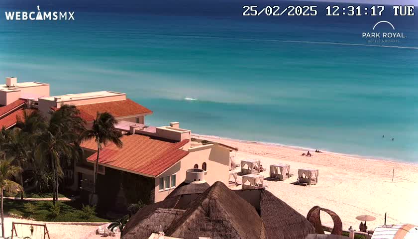 A beachfront resort with red-tiled roofs and palm trees overlooks a calm, turquoise ocean and a sandy beach on a sunny day.